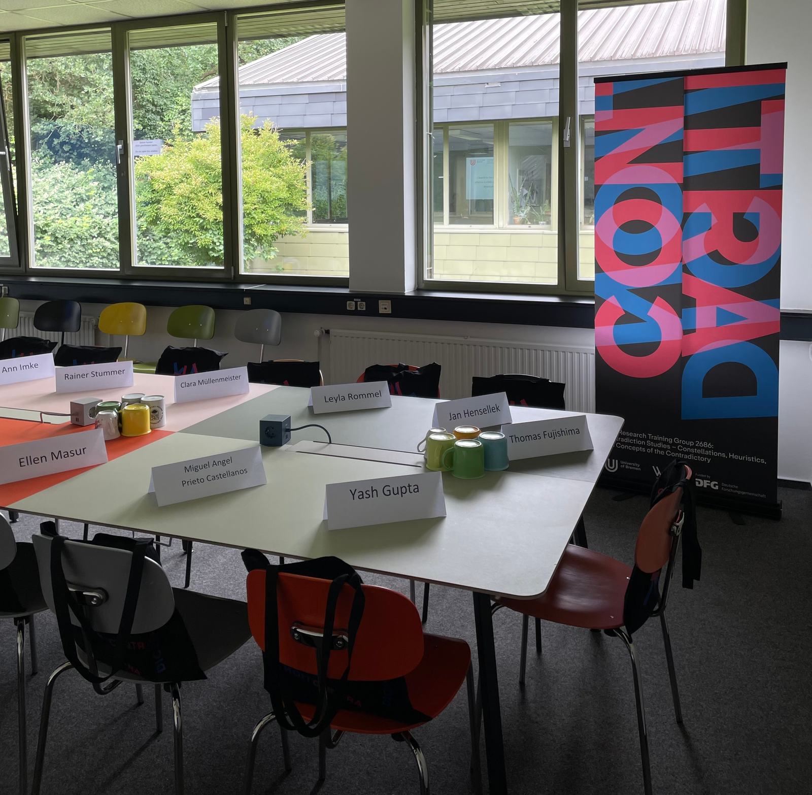 Zu sehen ist der Tisch in einem Büroraum des GRK Contradiction Studies. Auf dem Tisch sind Papierschilder mit den Namen der neuen Doktorand:innen und Welcome-Bags auf den Stühlen.